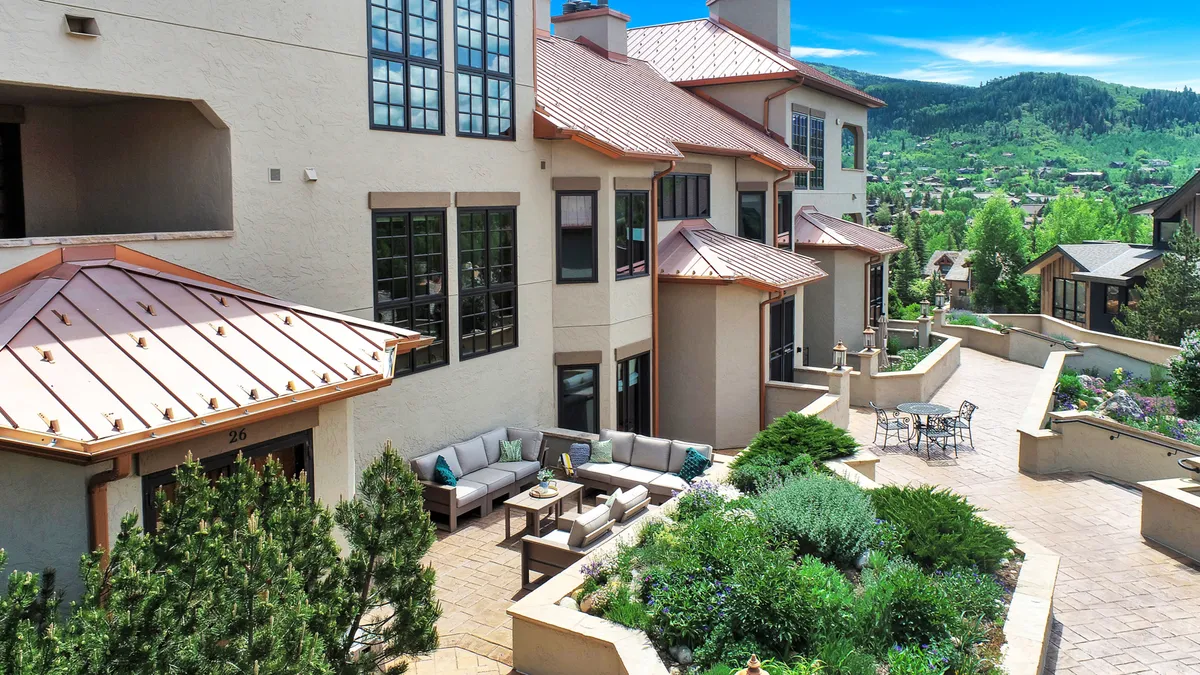This image features a patio seating area set among a landscaped garden, with the building's architecture visible and mountains in the distance. The outdoor setting is comfortable and welcoming, perfect for relaxing, enjoying coffee, or gathering with friends while taking in scenic views.