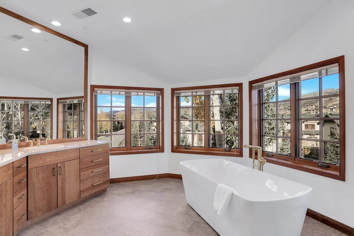 A luxurious bathroom with a freestanding soaking tub, large windows framing scenic outdoor views, wood cabinetry, and a broad vanity with mirrors. The room is bathed in natural light, offering a spa-like retreat for relaxation and self-care.