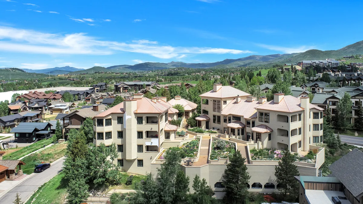 This image shows the exterior of the multi-level building surrounded by lush greenery, with the scenic mountain backdrop and the nearby residential area visible. The scene is welcoming, set in the golden hour of sunset, and hints at the relaxed, community-oriented atmosphere.