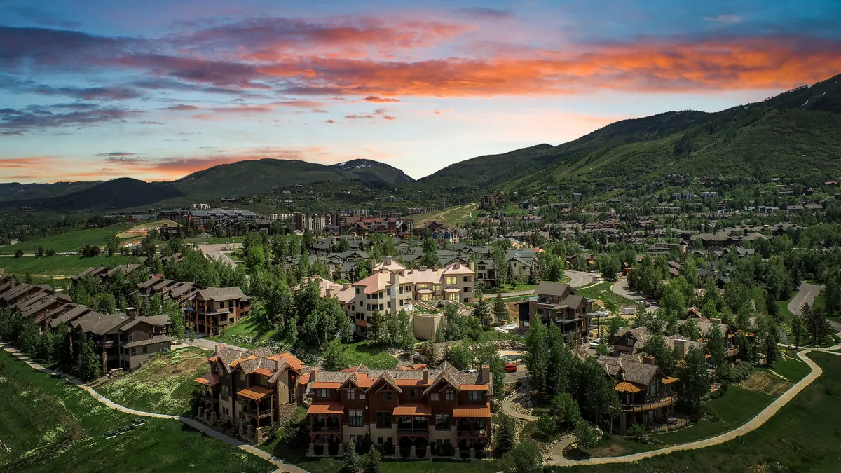 This image captures a sweeping view of the Steamboat Springs mountain range at sunset, with residential buildings nestled among lush greenery. The colorful sky and prominent natural setting evoke the year-round beauty of the area, highlighting both adventure and tranquility.