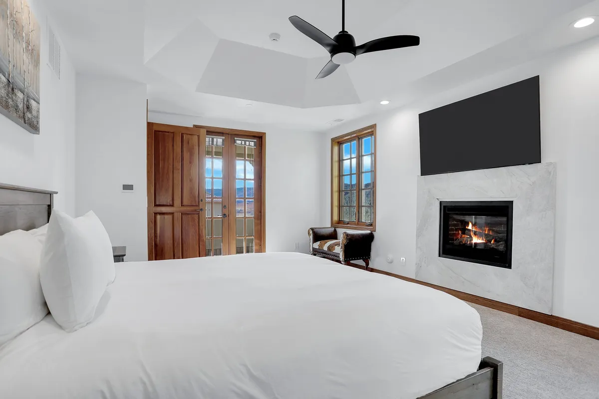 A serene bedroom featuring a king-sized bed with plush bedding, a cozy fireplace, large windows letting in abundant natural light, and wooden doors leading outside. The space exudes tranquility and comfort, ideal for restful nights and rejuvenating mornings.